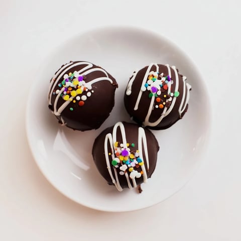 Seven glossy Hot Chocolate Bombs sit on a parchment-lined tray, topped with white chocolate drizzle and colorful sprinkles for a festive look.