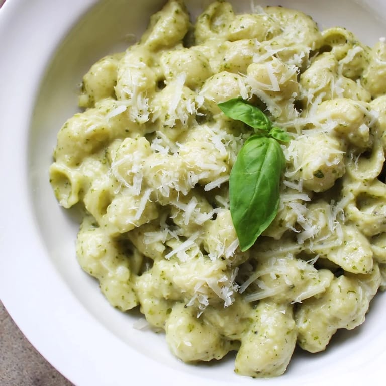 A close-up of Creamy Avocado Pesto Mac reveals rich green sauce clinging to tender pasta shells, suggesting a luscious, velvety texture.
